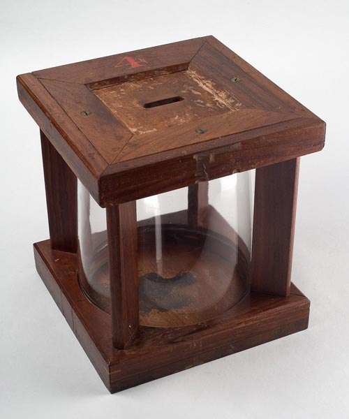 A nineteenth-century ballot box: a wooden frame around a clear glass jar, with a narrow slot cut in the lid