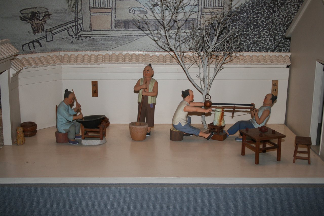 A museum diorama of a workshop: small modelled figures at low benches under a tiled roof, with a painted scene behind them