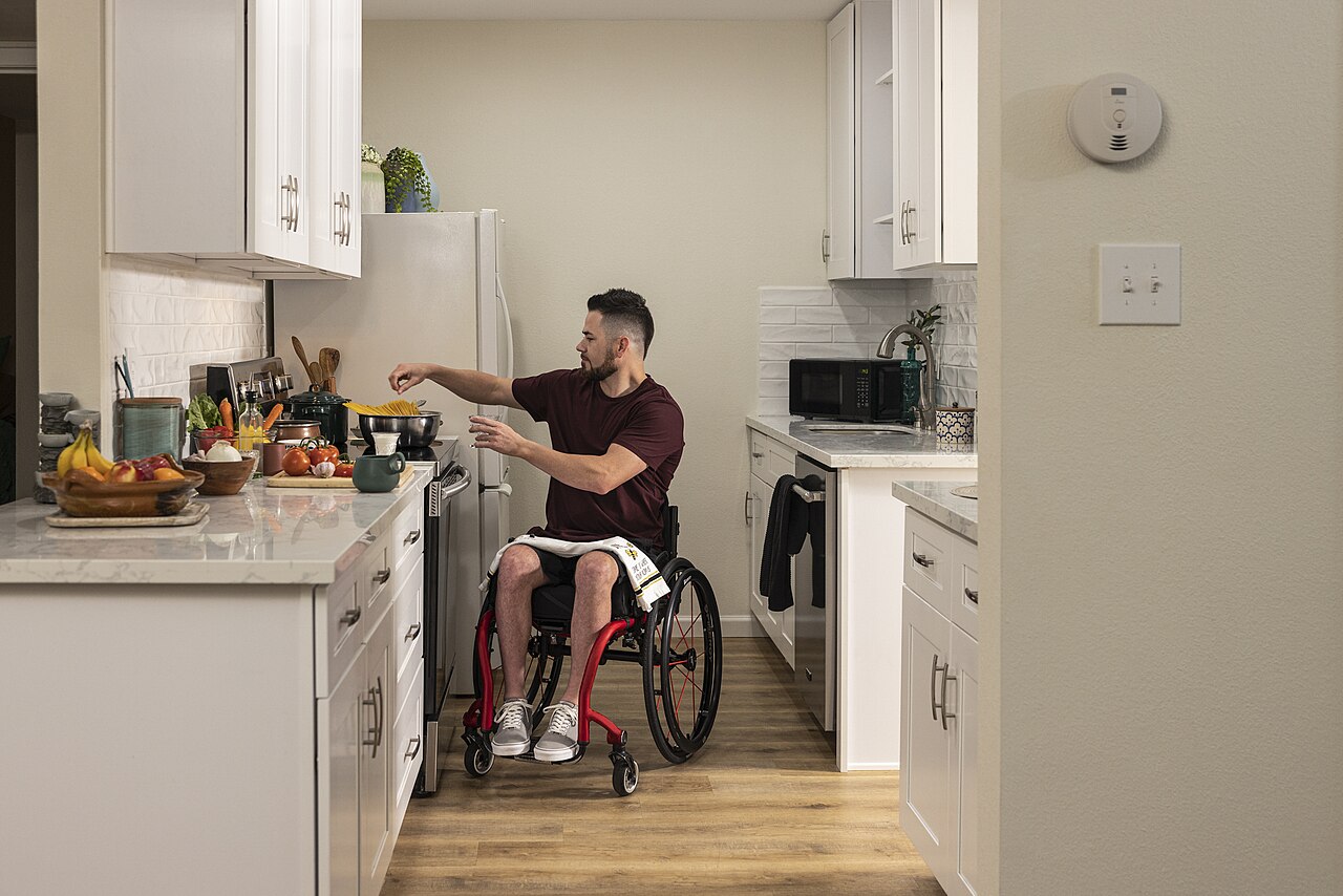 A man seated in a wheelchair at a kitchen counter, reaching over to stir a pan on the stove, with fruit and vegetables on the worktop beside him