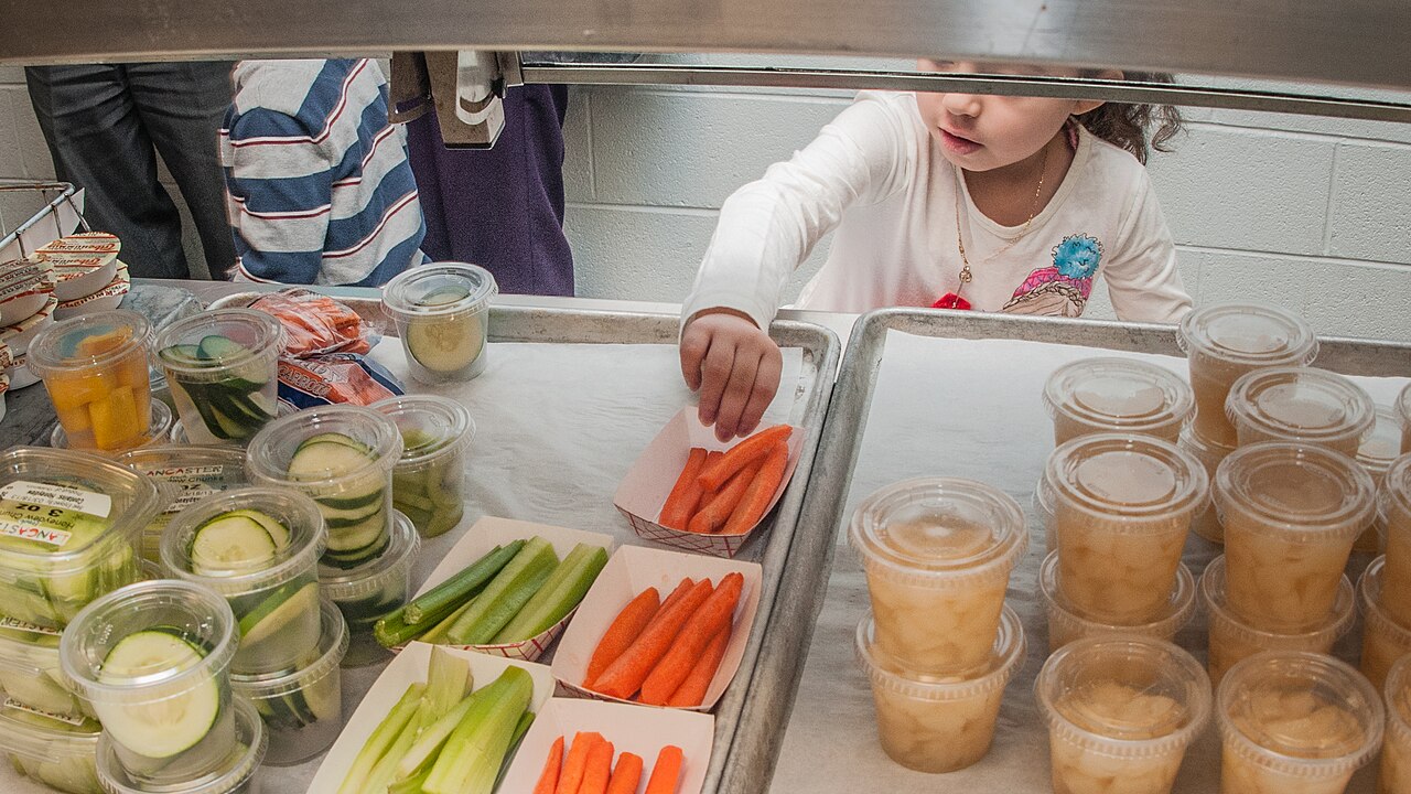 A school cafeteria serving counter seen from behind the food: a girl reaches across for a tray of carrot sticks, with cups of fruit and packets of cut vegetables laid out in front of her