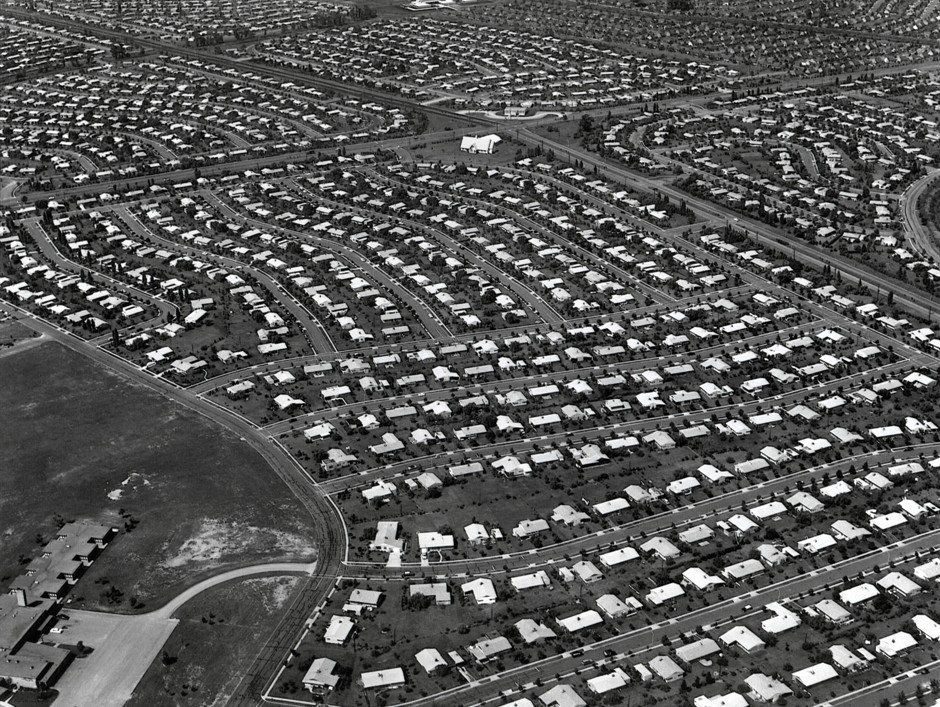 A black and white aerial photograph of a large postwar housing development, hundreds of near-identical single-storey houses laid out along curving streets with small matching yards