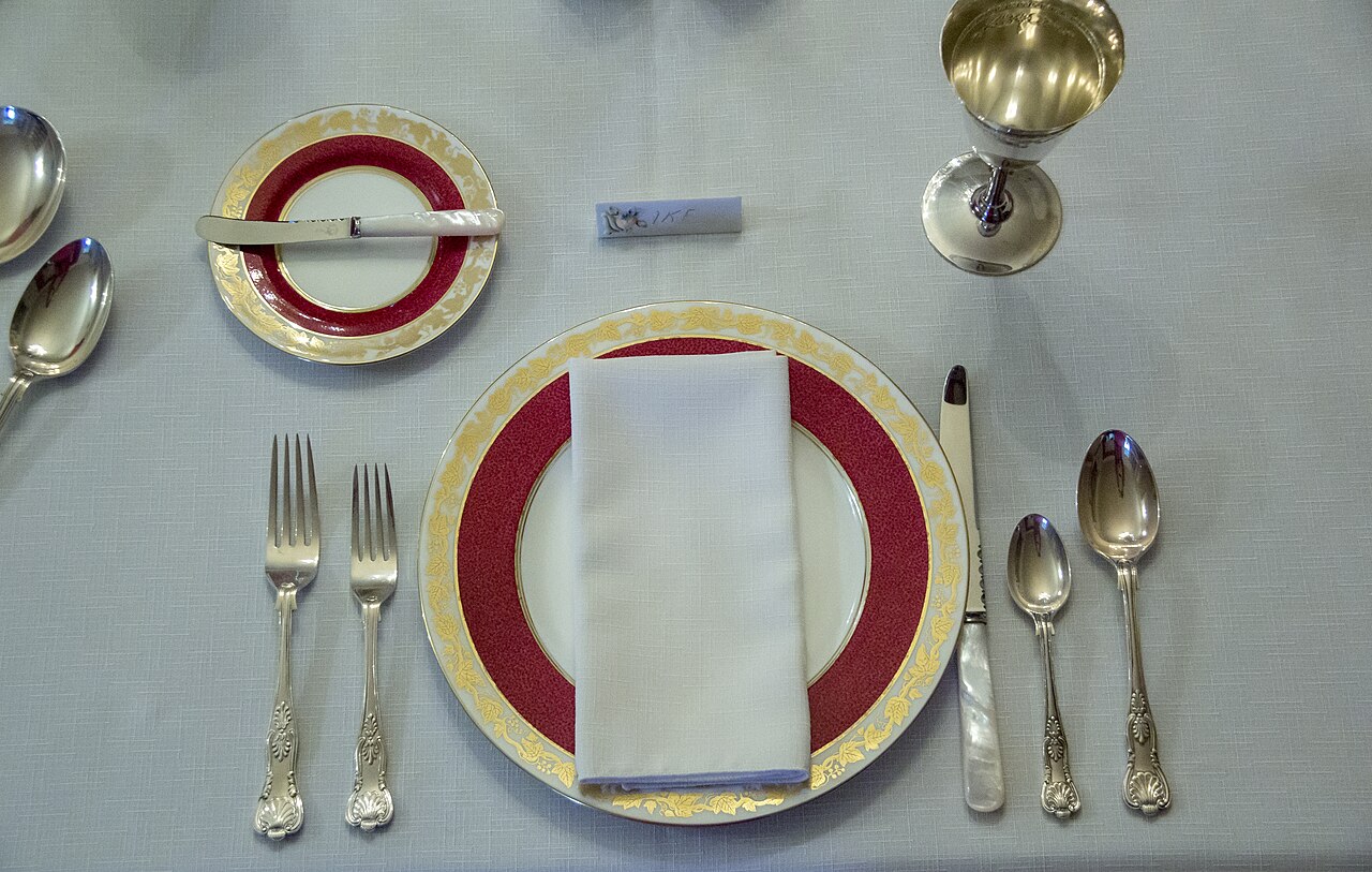 A long dining table set for a formal meal, with matching plates, several pieces of cutlery at each place, folded napkins, glasses and a low centerpiece running down the middle