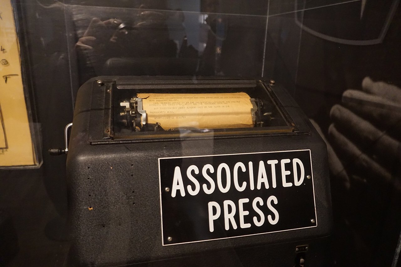 A black teletype machine in a museum case, a strip of yellowed paper with typed lines still in its printer, and a white-lettered sign on its front reading Associated Press