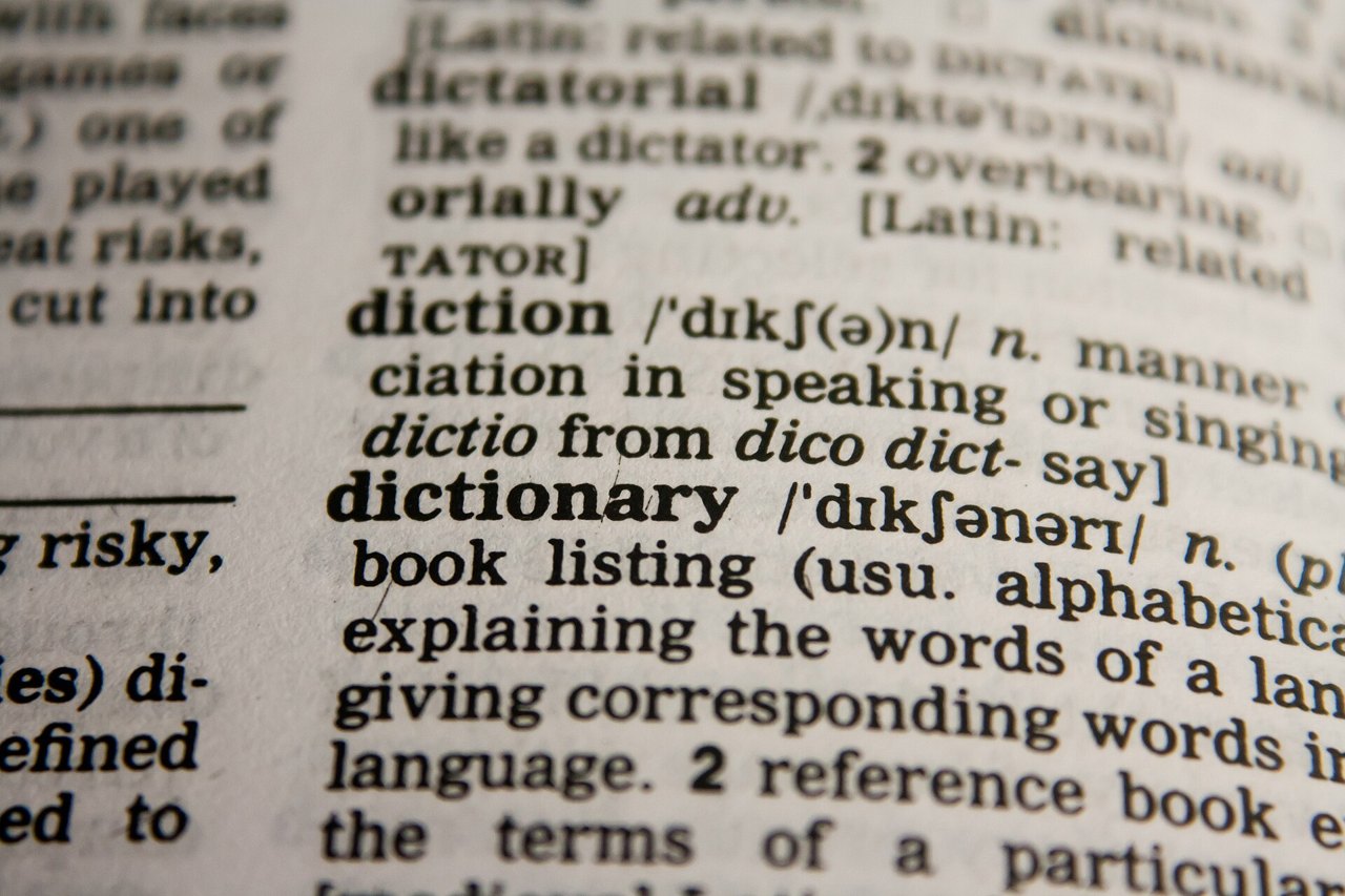 A close-up of a printed dictionary page in sharp focus on the entry for the word dictionary, with the entries for diction and dictatorial above it