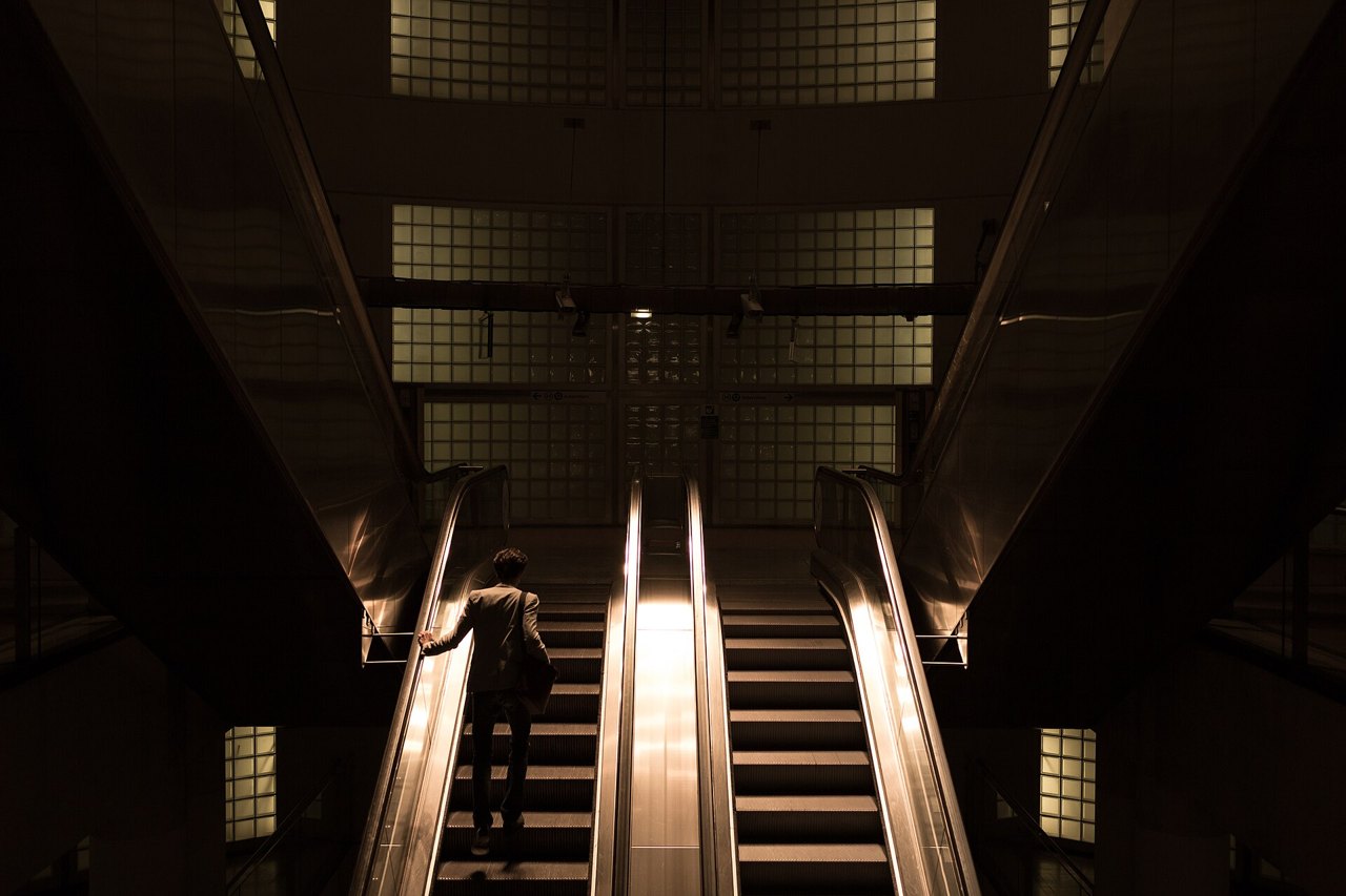 A dimly lit station hall with three moving staircases rising side by side toward glowing glass-block walls, a single man in a coat riding the left one