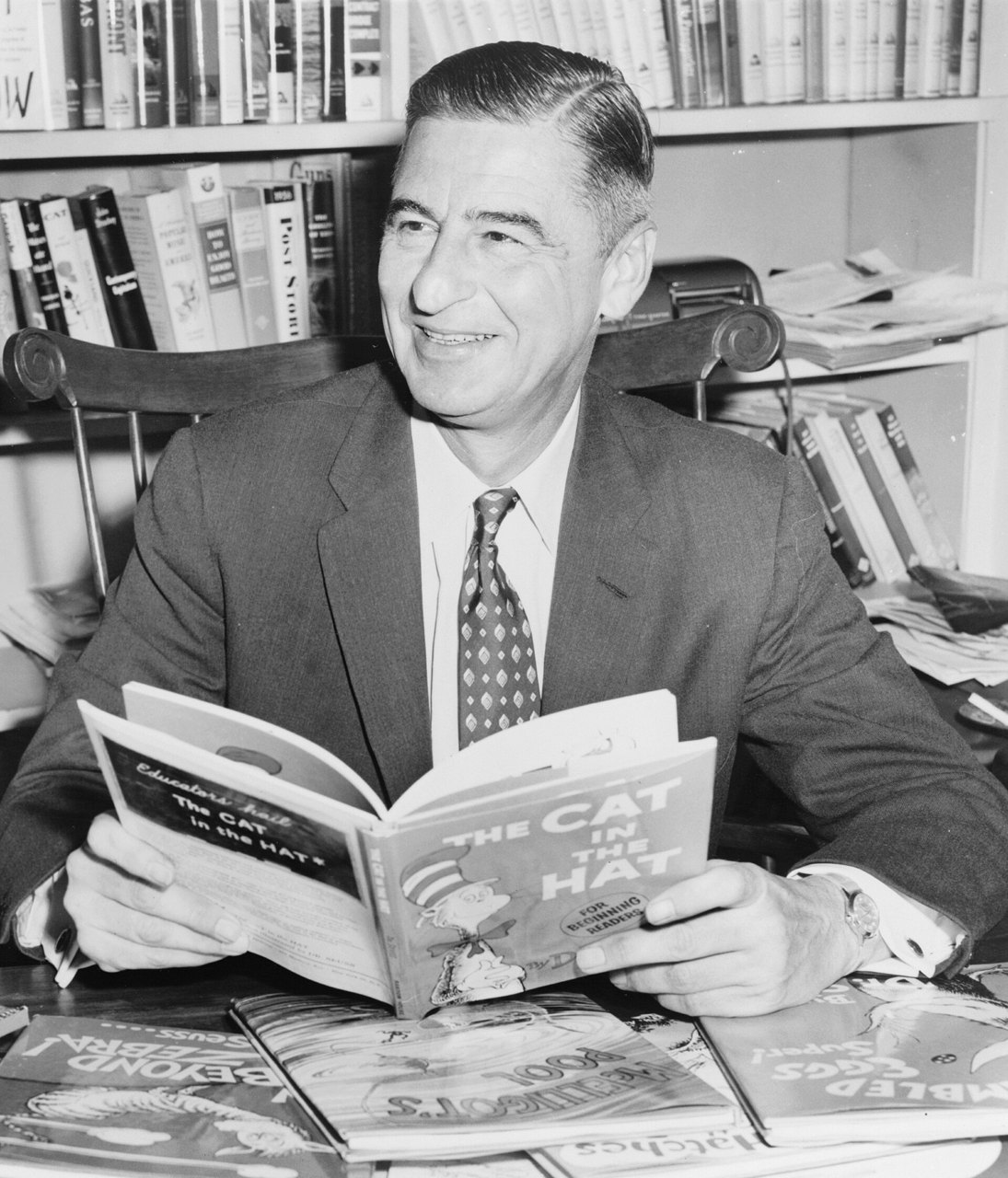 A black-and-white photograph of a smiling man in a suit and patterned tie seated at a desk in front of bookshelves, holding open a copy of The Cat in the Hat