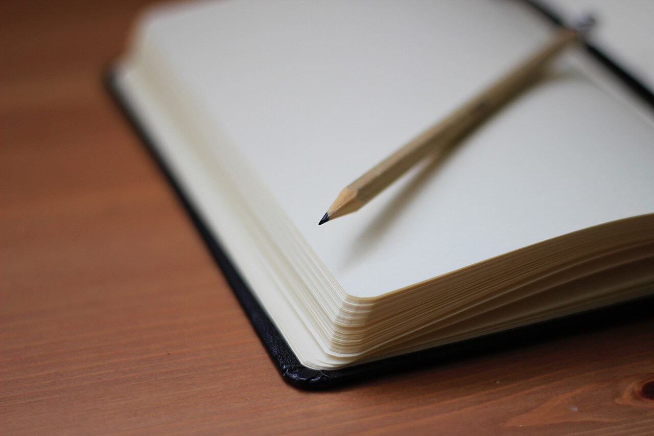 A blank notebook lying open on a wooden table, its cream pages empty, with a sharpened wooden pencil resting across the right-hand page