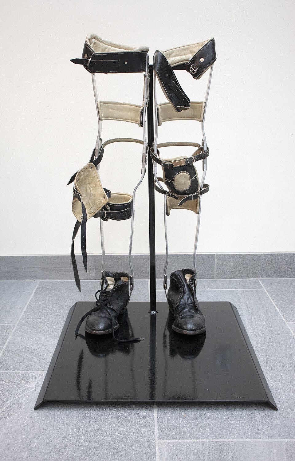 A pair of child-size metal leg braces with leather straps and attached black leather boots, displayed on a stand in front of a white wall