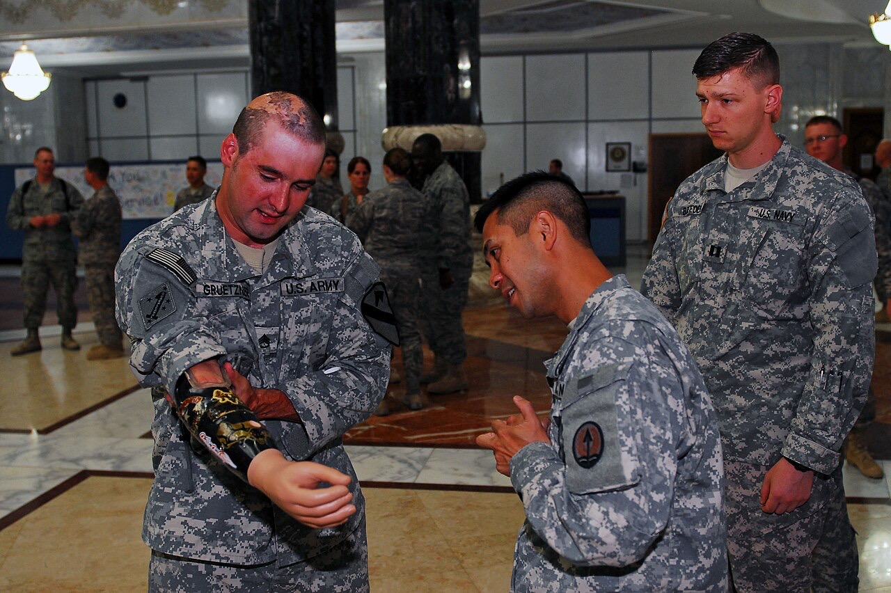 A U.S. Army veteran in uniform adjusts the decorated prosthetic forearm and lifelike hand he wears, while two soldiers look on in a marble-floored hall