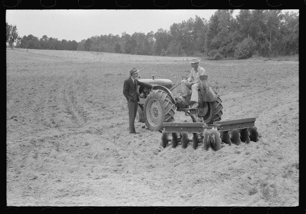 A black-and-white 1940 photograph of a farmer seated on a small tractor pulling a disc plow across a bare field while another man stands beside it talking to him
