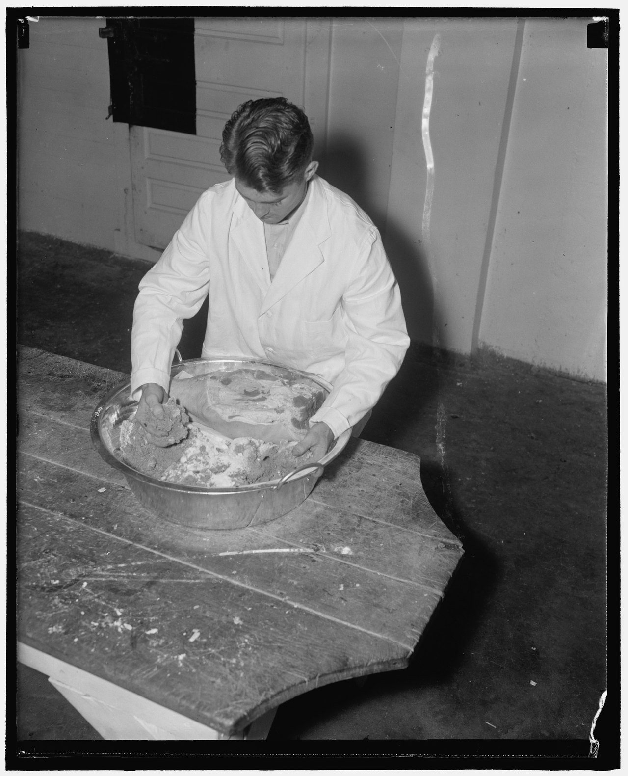 A black-and-white 1938 photograph of a man in a white coat bending over a table, rubbing a large ham in a basin with his hands