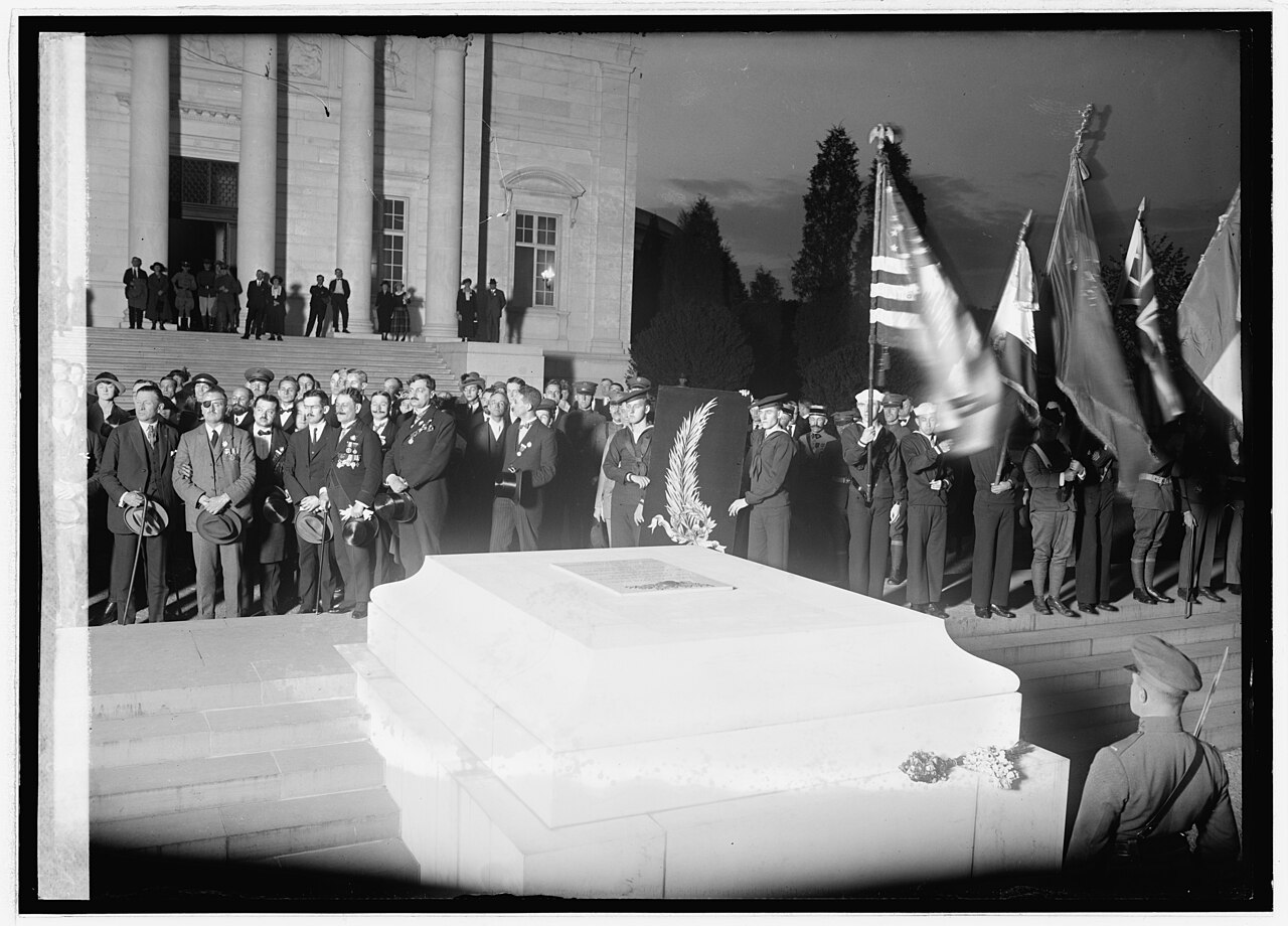 A large crowd in coats and hats standing in rows on open ground at a cemetery, facing a low stone monument, with bare trees behind them
