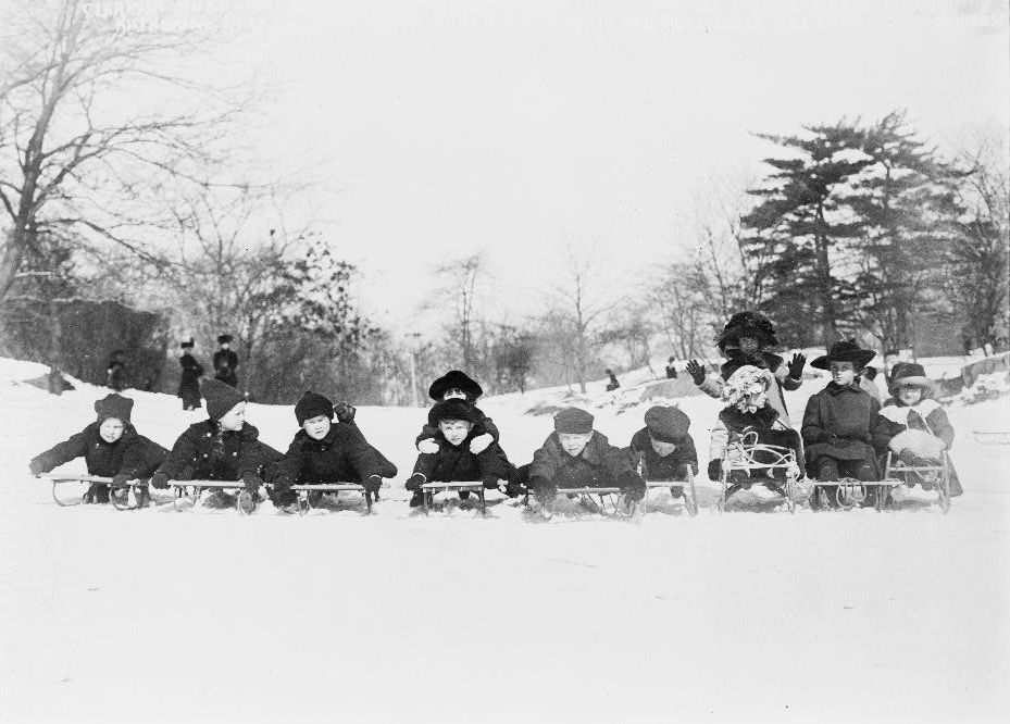 An old photograph of a row of children lying on wooden sleds in the snow, lined up side by side and looking at the camera, with an adult standing behind them waving