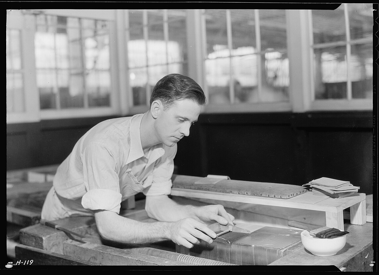 A man in shirtsleeves bends over a workbench in a printing works, laying gold leaf onto the spine of a book by hand, with tools and stacked work around him