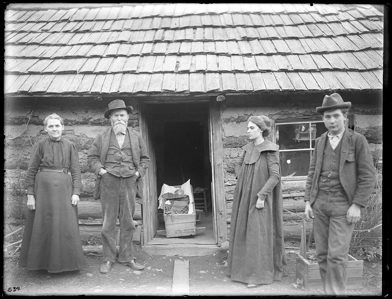 A family standing and seated outside a log cabin: a young couple, an older man and woman, and a baby in a homemade cradle on the ground in front of them