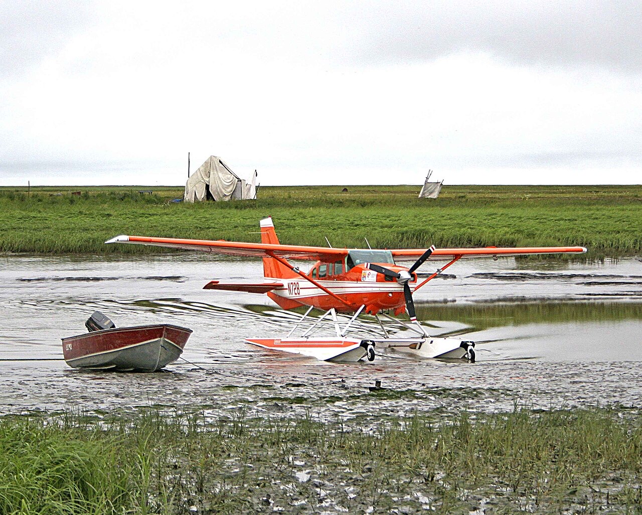 An orange-and-white single-engine floatplane resting in a muddy tundra river channel beside a small open boat, with canvas tents and flat green tundra stretching behind