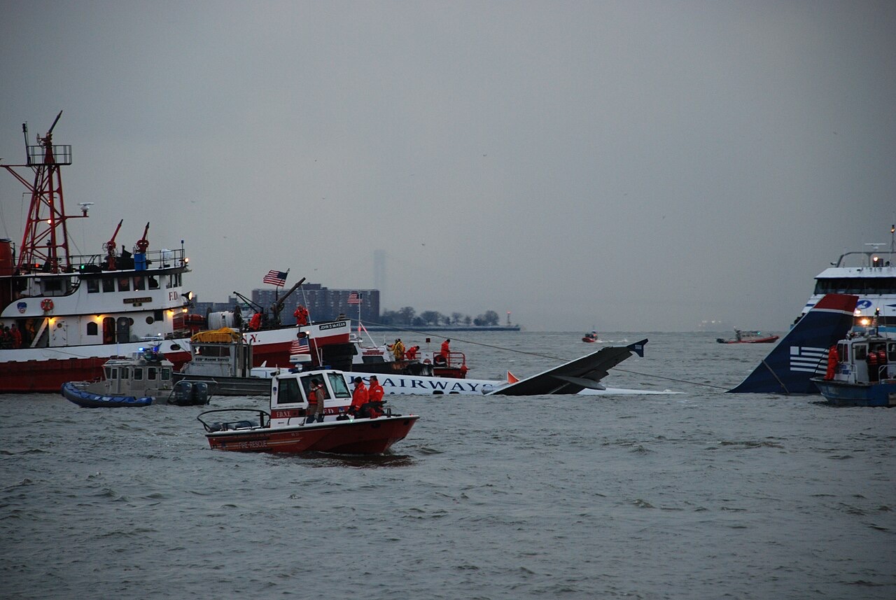 A partly sunken airliner floating low in a gray river, its tail and the words US Airways visible, surrounded by fireboats, a small red fire-rescue boat and a ferry