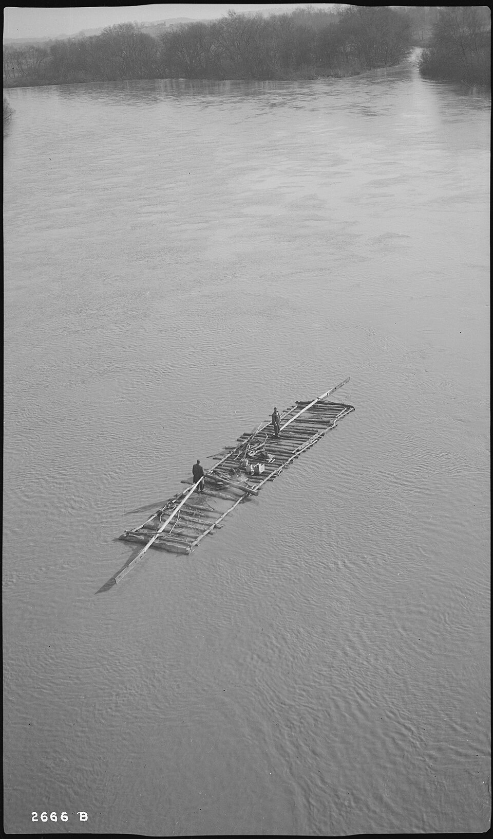 A black-and-white aerial photograph of two men standing on a long, narrow raft of lashed logs with long steering sweeps at each end, midstream on a wide, calm river