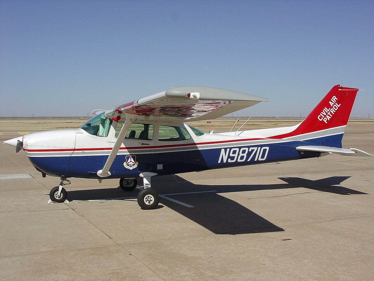 A red, white and blue Cessna 172 with Civil Air Patrol markings parked on a sunlit concrete flight line under a clear sky