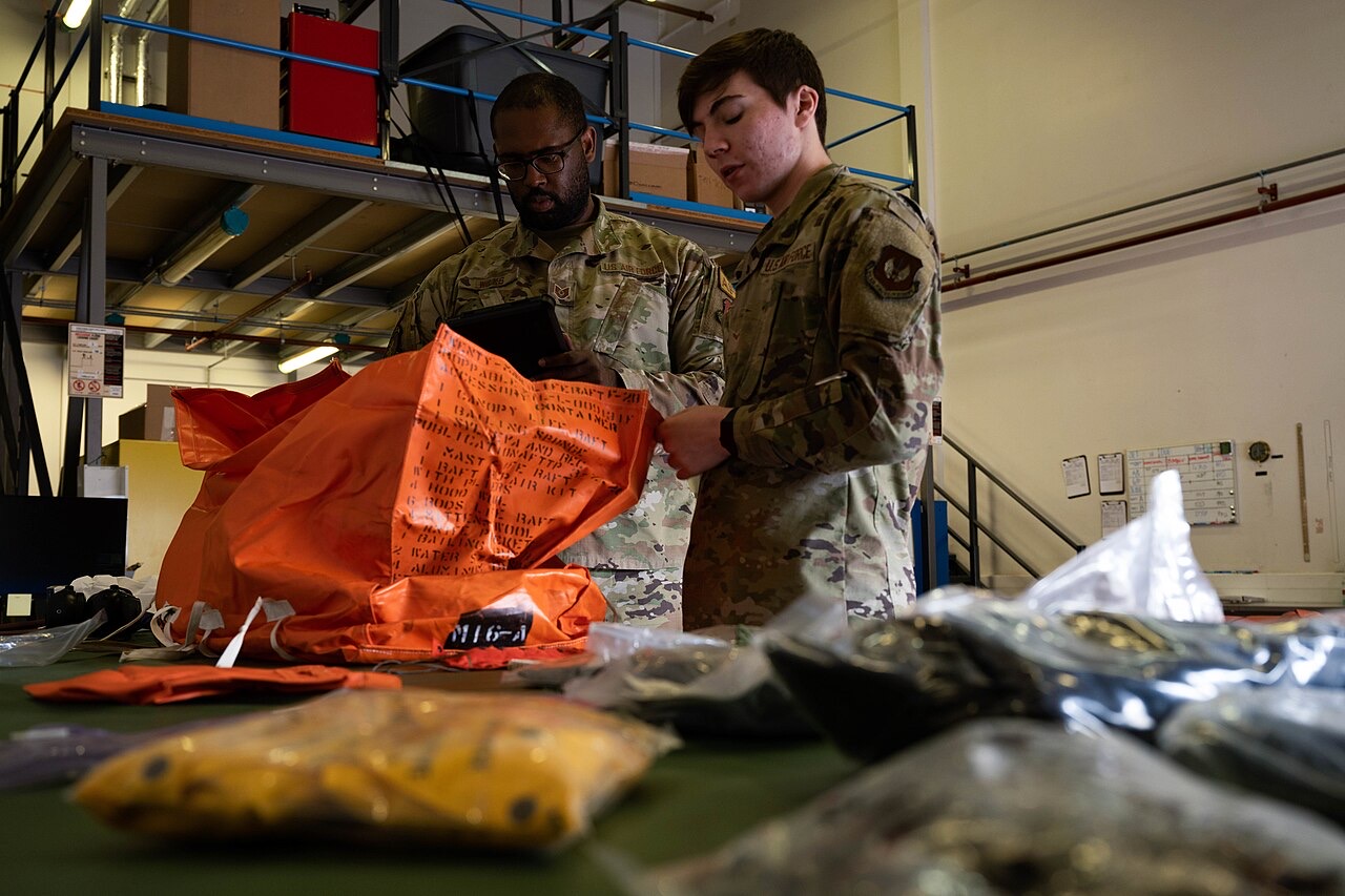 Two U.S. Air Force airmen at a table covered with packed survival equipment; one reads from a tablet while the other handles a bright orange life-raft bag stenciled with a packing list