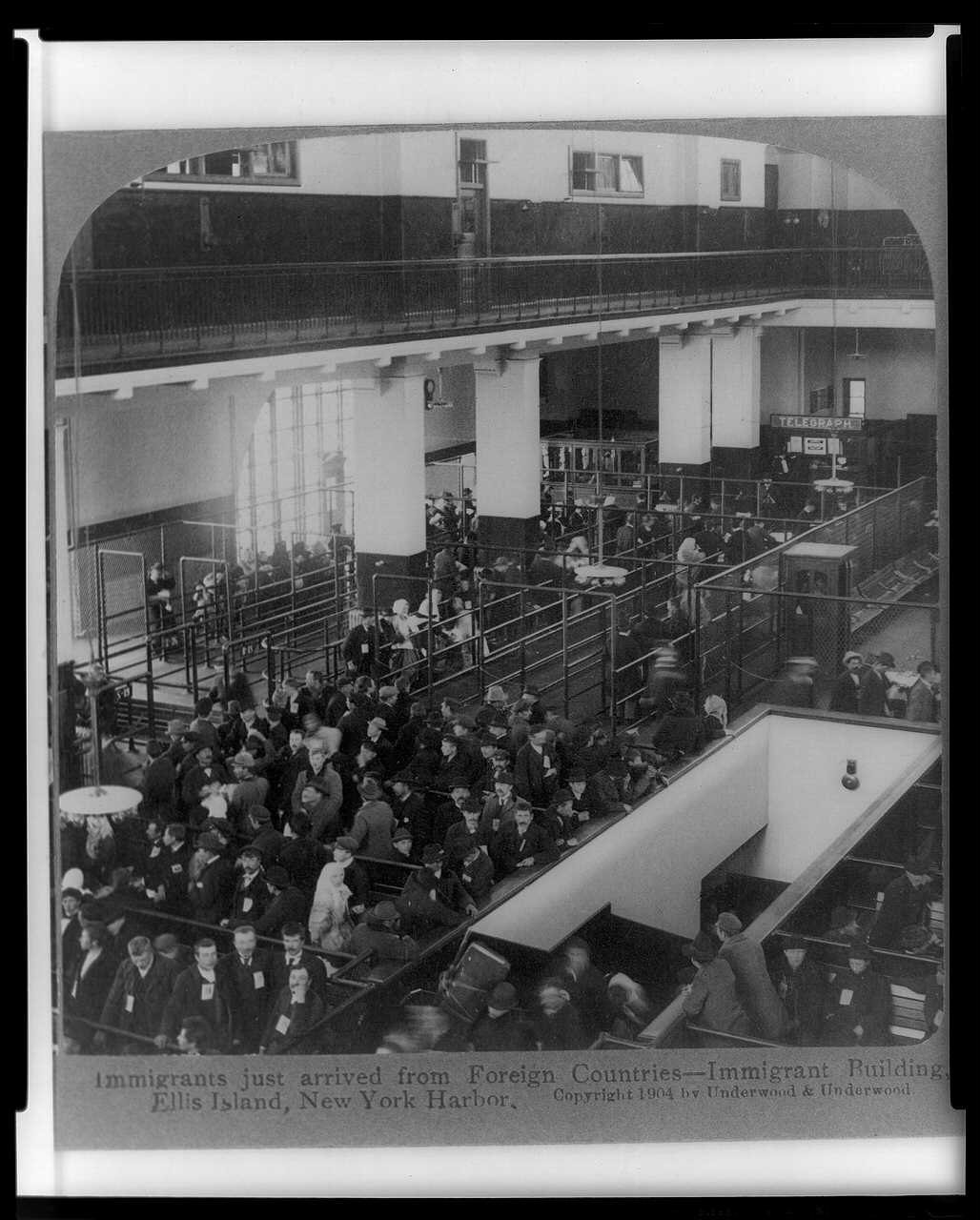 A black-and-white 1904 photograph of a large hall packed with newly arrived immigrants standing in railed lines beneath a balcony