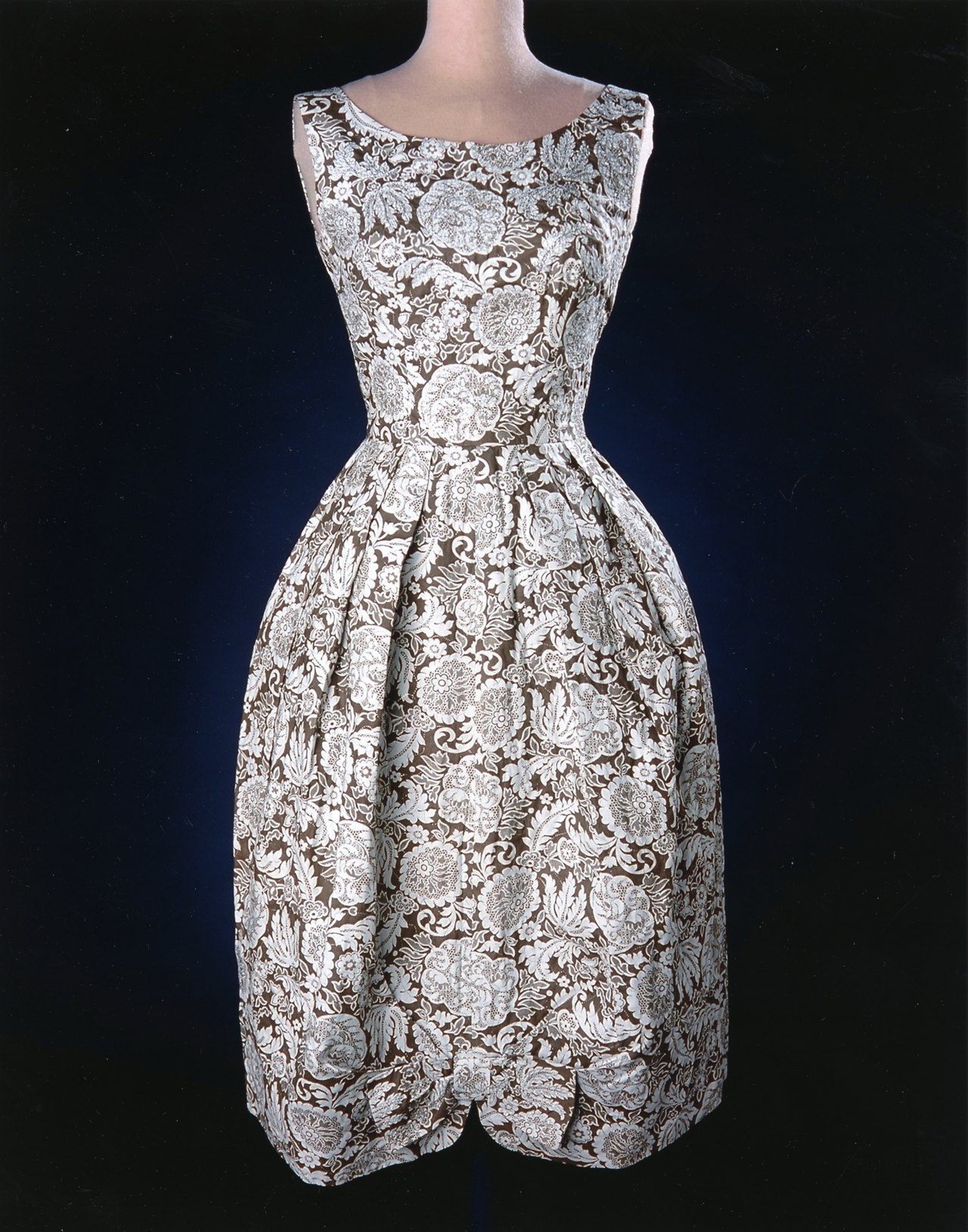 A sleeveless dress with a full skirt in a brown-and-white floral print, displayed on a mannequin against a dark background