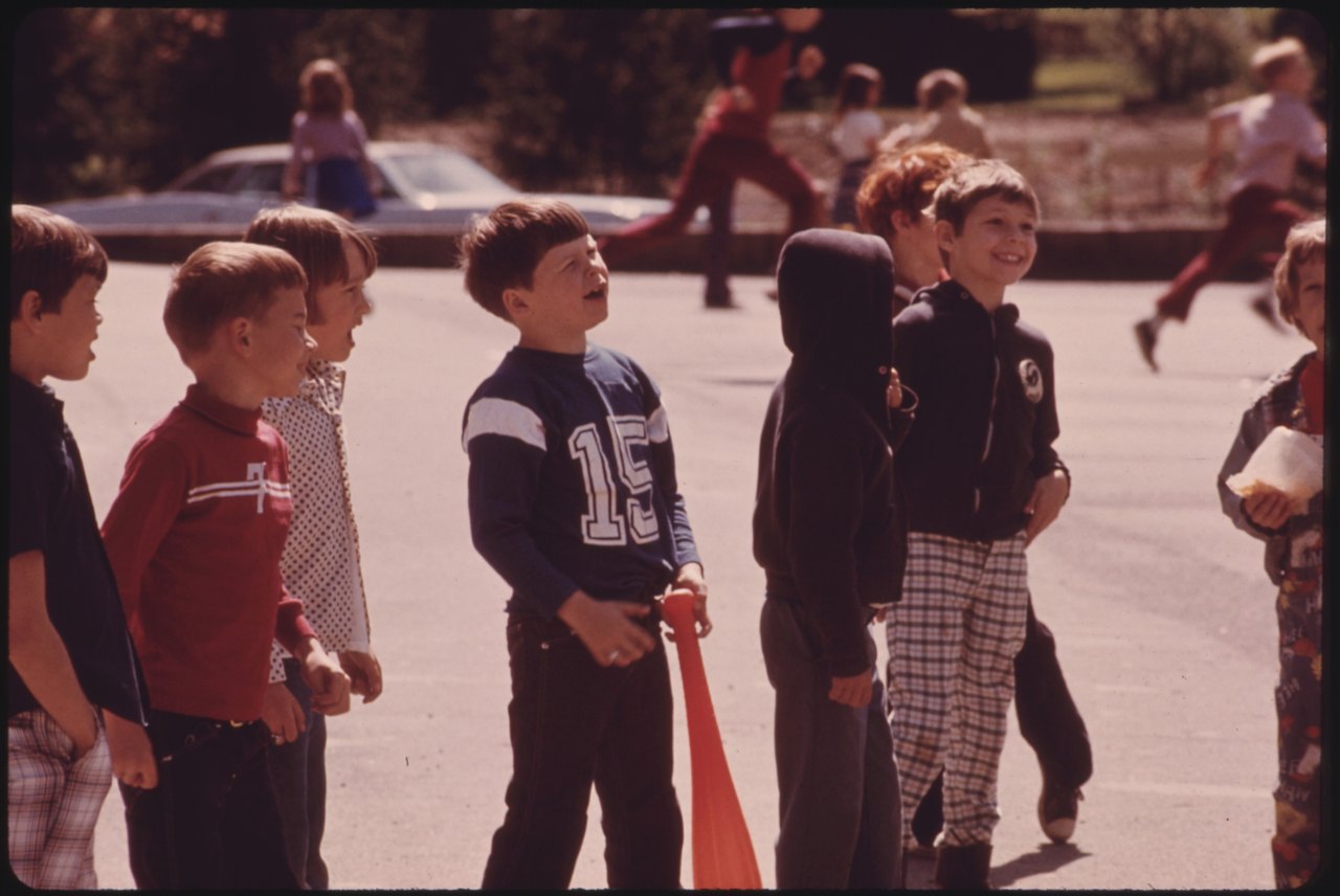 A color 1970s photograph of a cluster of boys in a paved schoolyard, several laughing and looking the same way, with other children and cars behind them