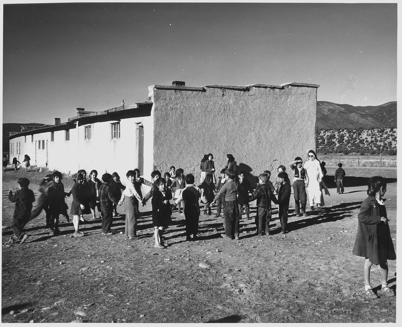 A black-and-white photograph of children holding hands in a ring game on a bare schoolyard beside a whitewashed adobe school, with hills behind