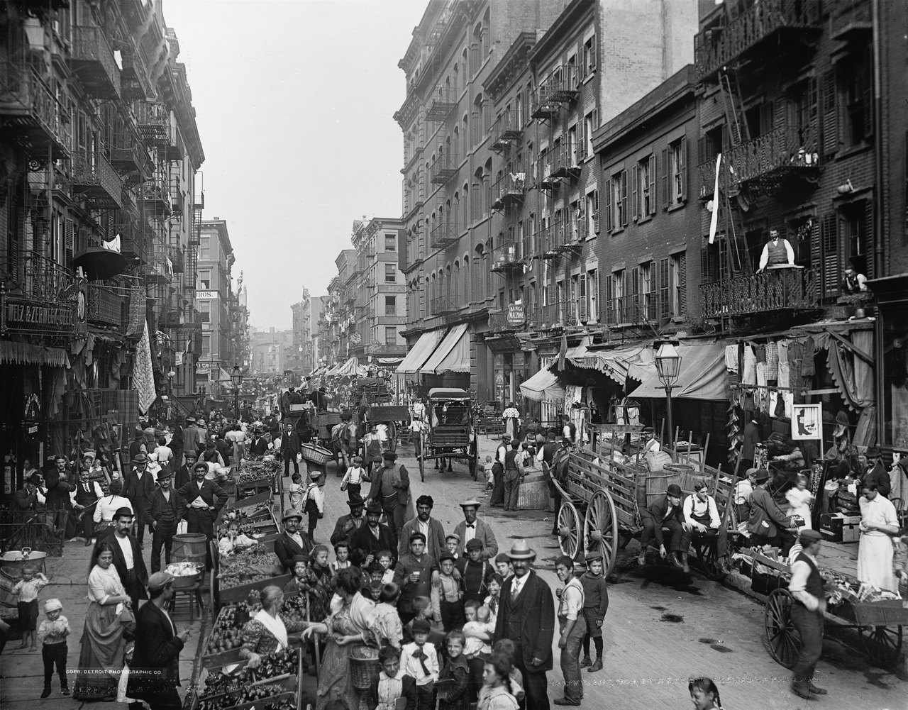 A hand-colored photograph of a crowded city street lined with tall tenements, pushcarts, awnings, horse-drawn wagons and hundreds of people