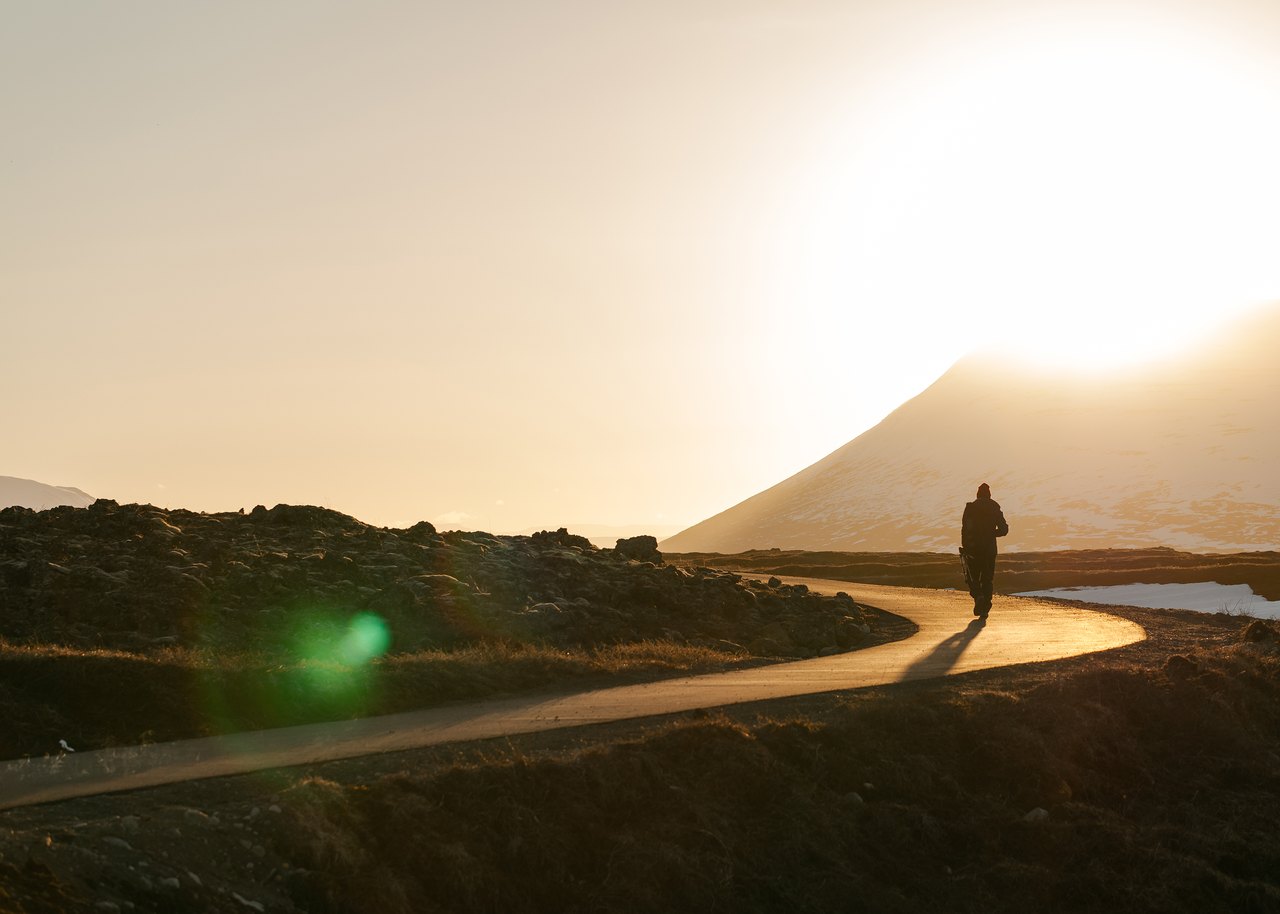 A lone figure walks away along a narrow road that curves toward a hill, with the low sun flaring gold in a hazy sky