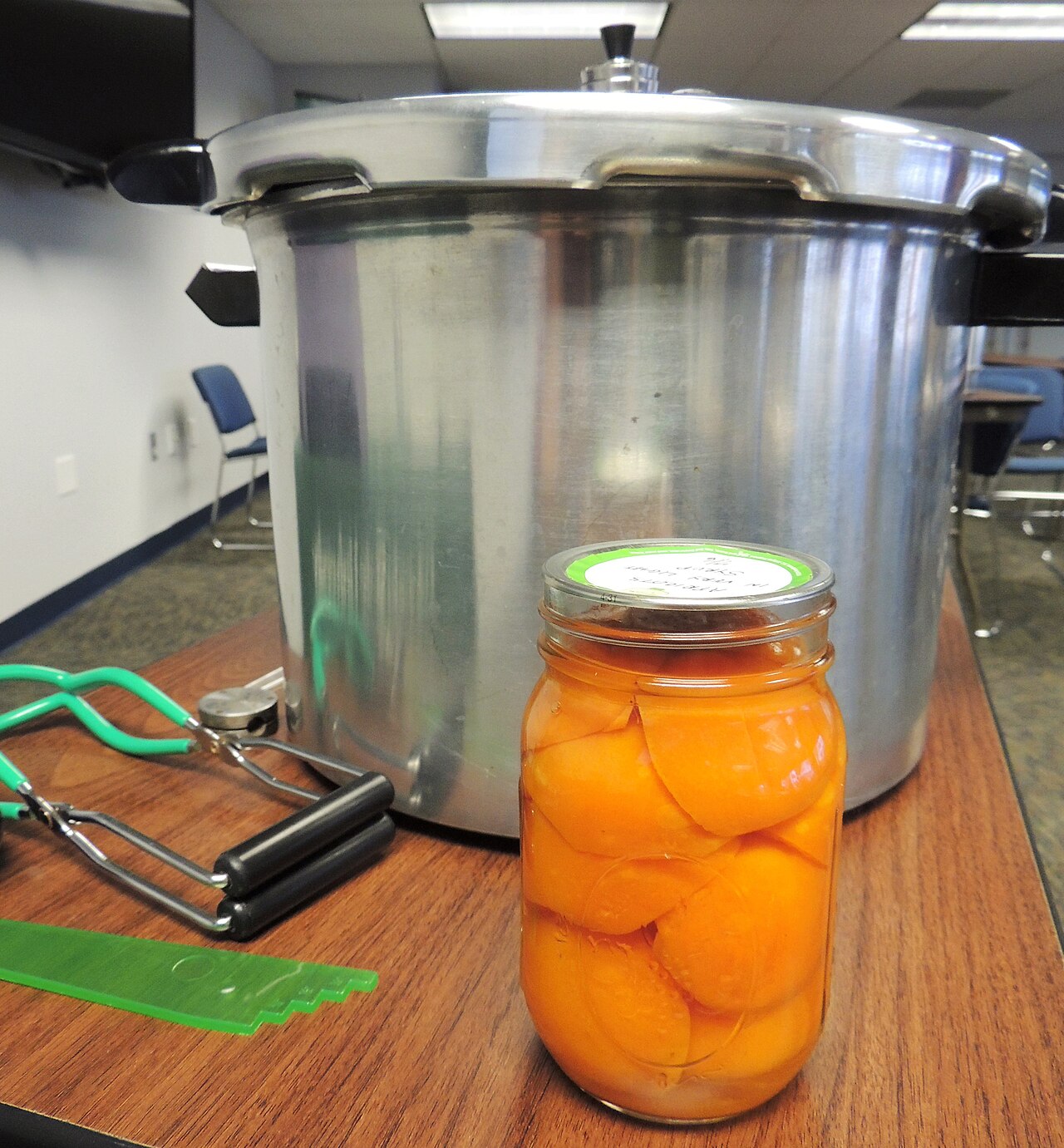 A sealed glass jar packed with peach halves in syrup, standing on a table in front of a large metal pressure canner