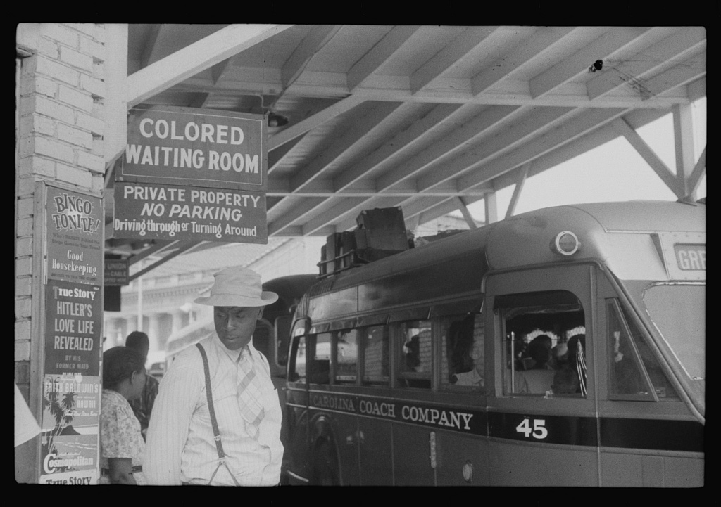 A black and white photograph at a bus station in 1940. A large hanging sign reads COLORED WAITING ROOM. A man in a hat and a woman stand beneath it beside a parked coach