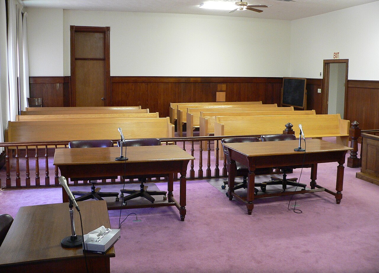 An empty American county courtroom with two wooden counsel tables and microphones in front of a low railing, with rows of public benches behind it
