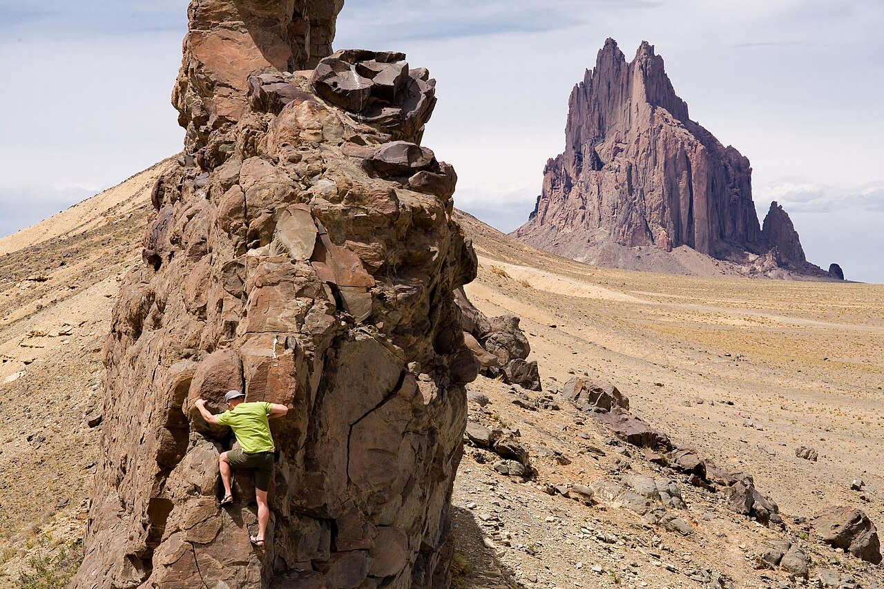 A tall jagged rock peak rising alone from a wide flat desert plain, with a person climbing a smaller rock ridge in the foreground