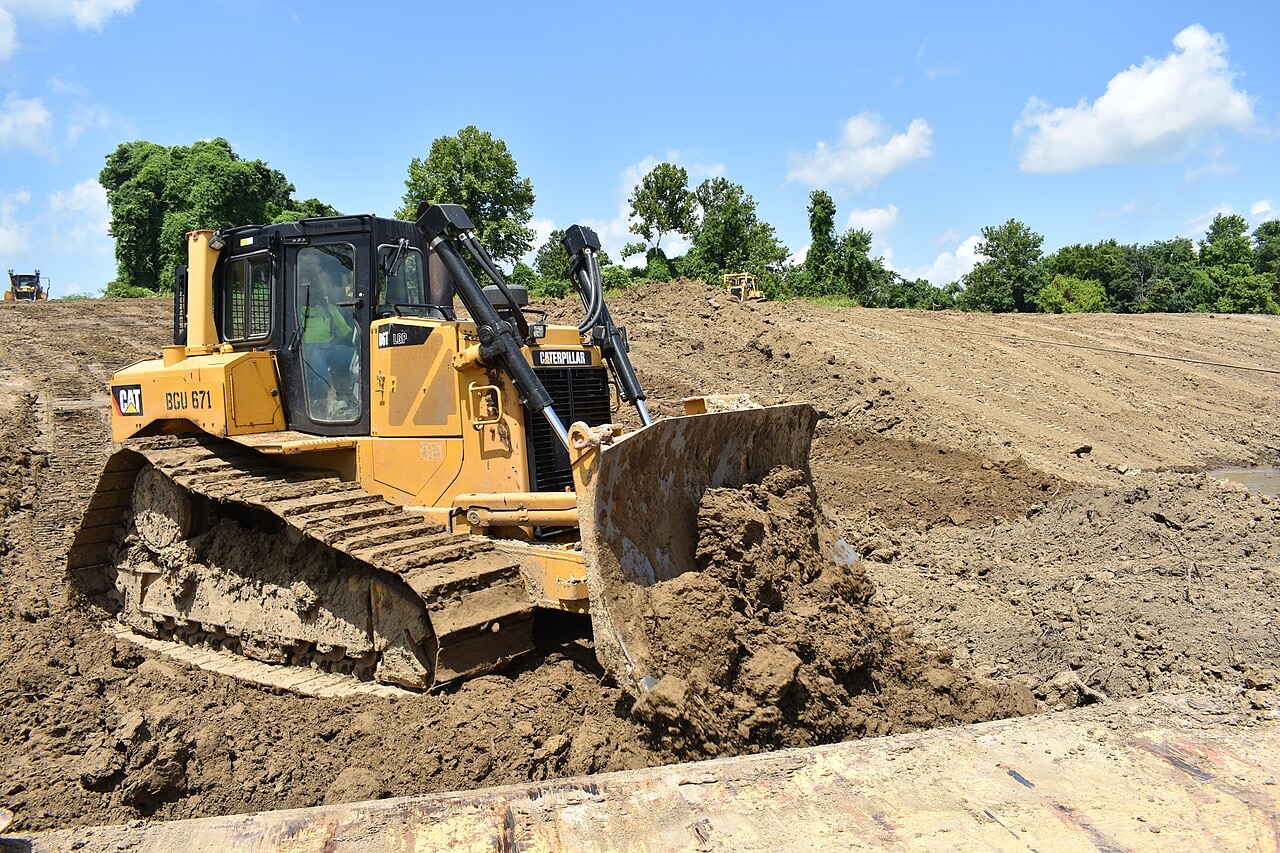 A yellow tracked bulldozer pushes a blade full of brown dirt across a wide slope of freshly graded bare earth, with green trees and blue sky behind