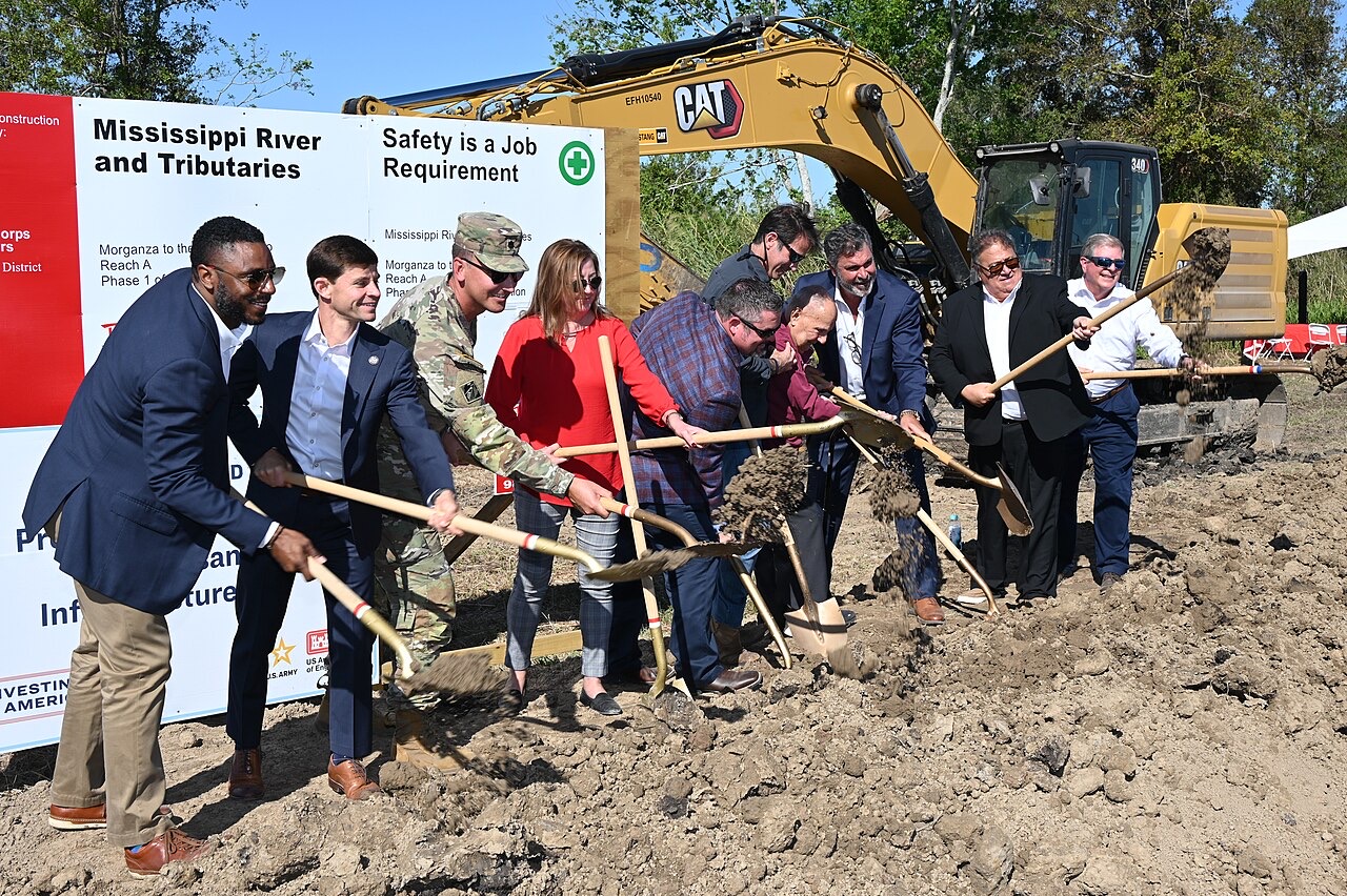 Officials in suits and one in camouflage uniform swing golden shovels of dirt in front of project signboards and a large yellow excavator