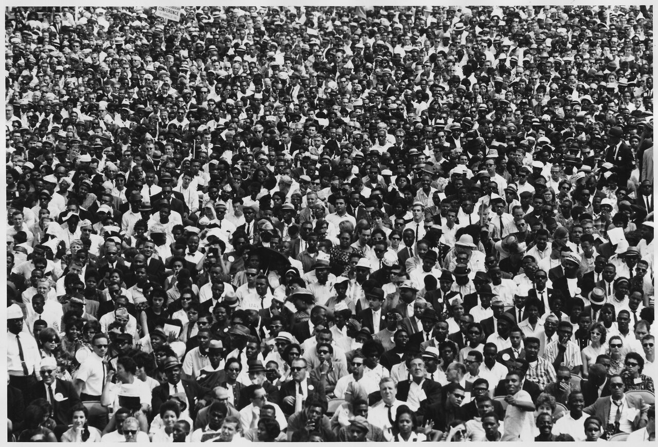 A black-and-white photograph of an enormous, orderly crowd of men and women in 1960s dress, filling the frame from edge to edge