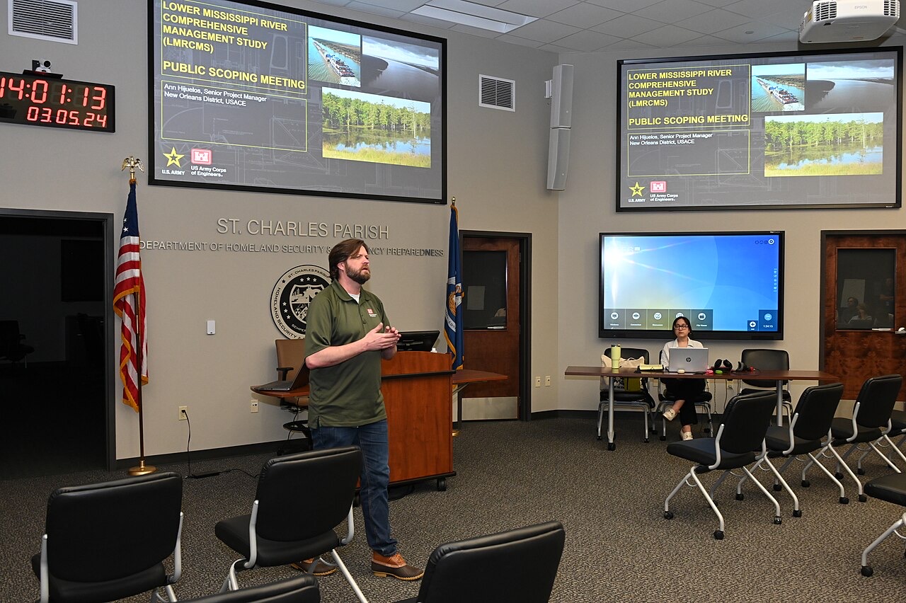 A government meeting room with rows of chairs, flags, and two large screens reading Public Scoping Meeting while a presenter in a green polo speaks