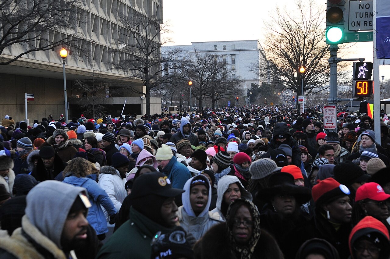 A dense crowd of people in winter coats and hats fills a city street between bare trees and stone buildings, stretching back as far as the eye can see