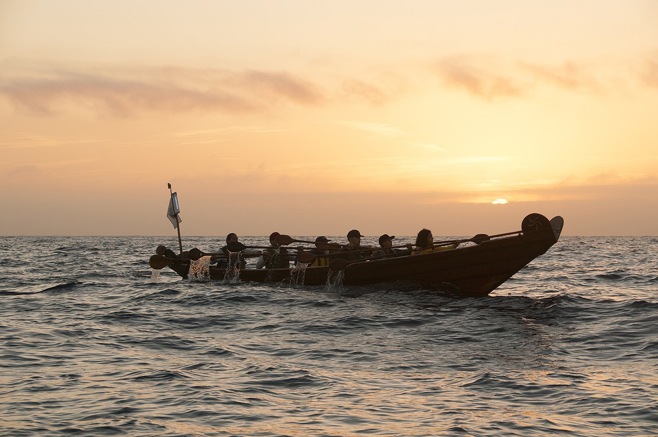 A long wooden plank canoe crowded with paddlers crossing open ocean at sunrise, its low hull silhouetted against an orange sky