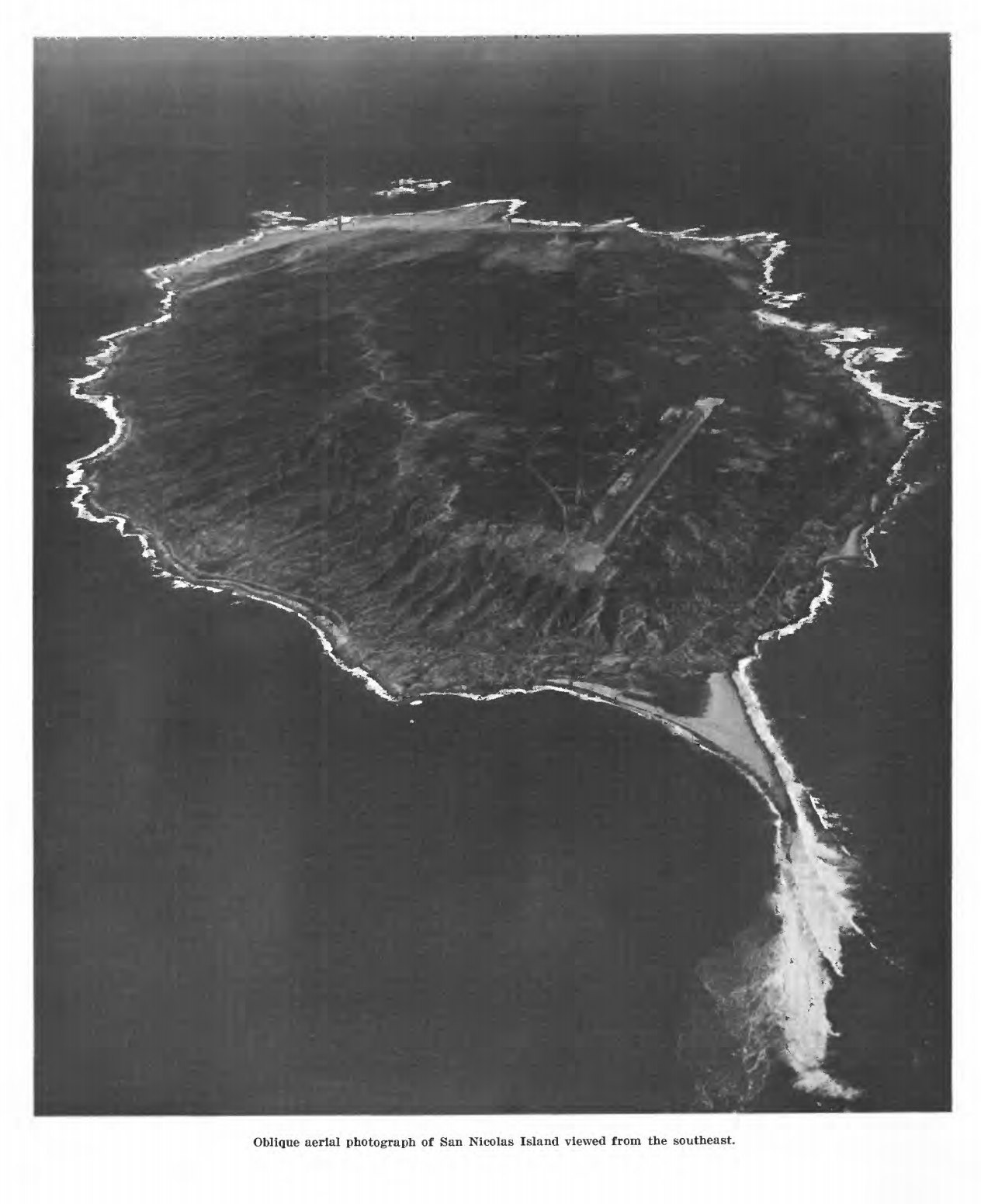 An oblique aerial photograph of a long flat island ringed by pale surf, its bare tableland falling away to cliffs and a spit of sand at one end