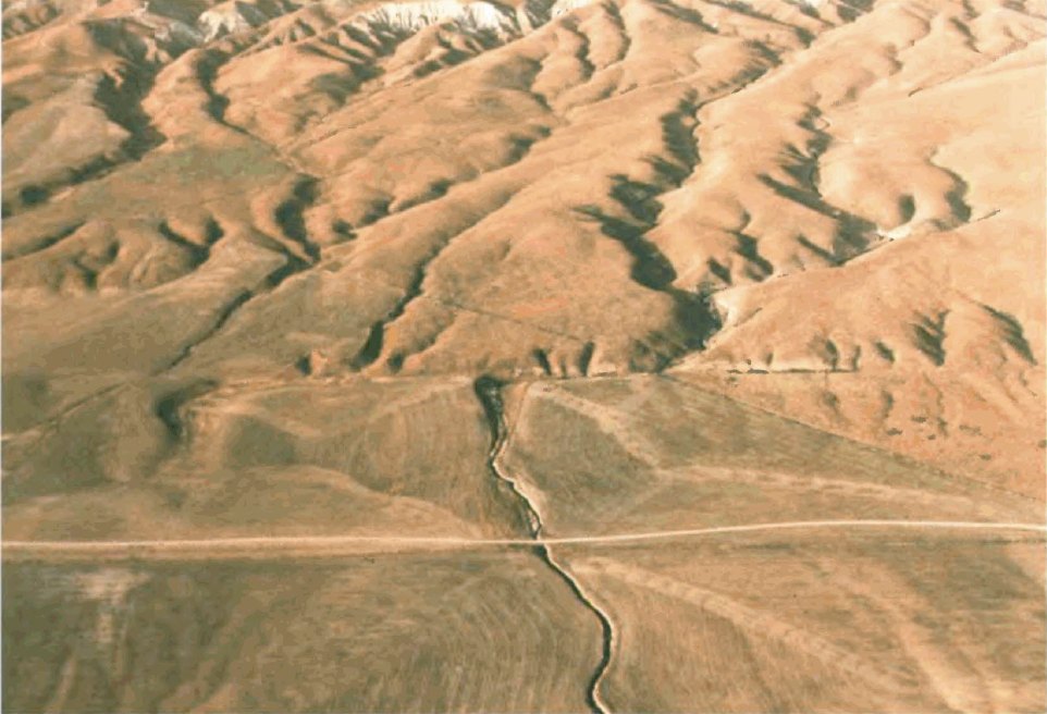 An aerial view of dry golden hills crossed by a long straight scar, where a dry streambed is bent sharply sideways where it meets the line