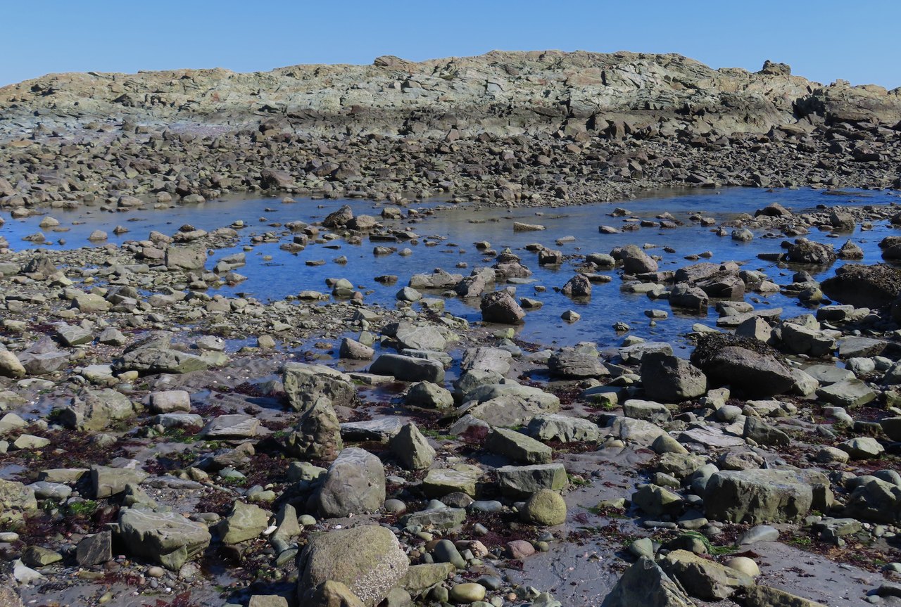 A wide rocky shore at low water, strewn with dark boulders, with shallow pools of trapped seawater reflecting the sky between them