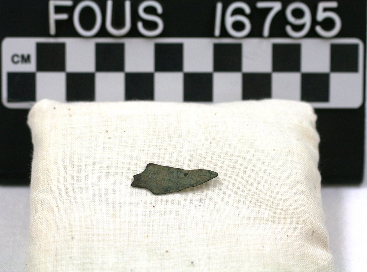 A small dark triangular projectile point resting on a white museum cushion beside a black-and-white checked scale bar marked in centimeters