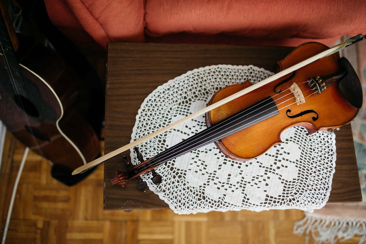 A violin lying on a small table with its bow across it, the four strings running down to the bridge, and the dark body of a cello standing behind