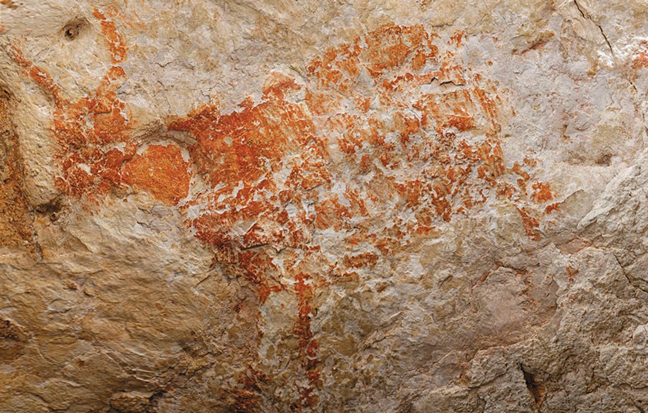 A large reddish-orange animal figure painted on rough pale cave rock, its body outlined and partly filled, with horns and legs clearly drawn