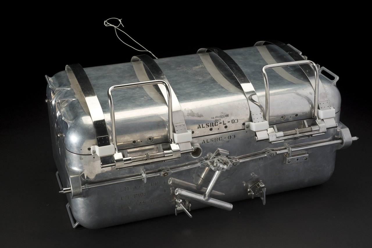 A rectangular aluminum box with metal straps, latches and a lifting handle, photographed against a black background