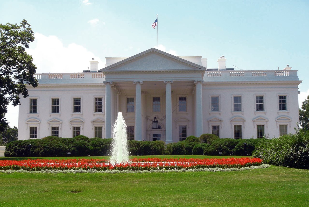 The white columned north front of the White House under a blue sky, with a fountain and red flowerbeds on the green lawn in front