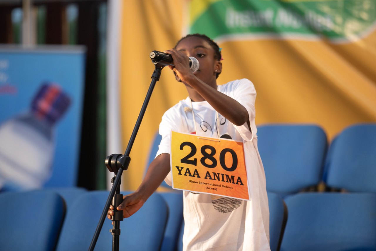 A young girl stands at a microphone on a stage wearing a numbered contestant card, one hand raised as she spells a word aloud to the judges
