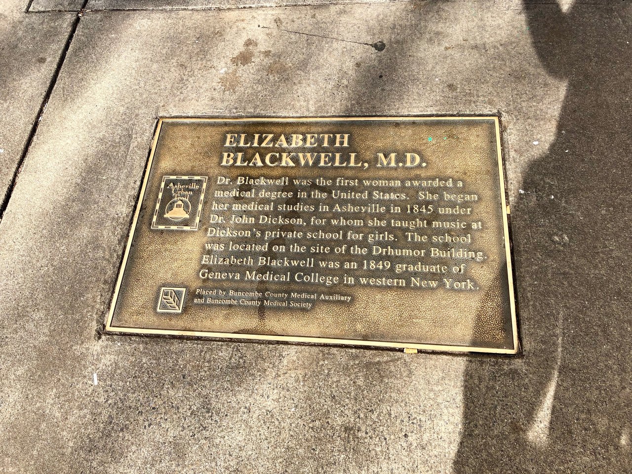 A metal historical marker on a post beside a street, its raised lettering naming Doctor Elizabeth Blackwell, with trees and buildings behind